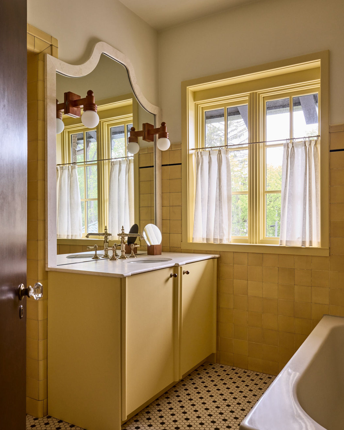 A vintage-style bathroom interior with mustard yellow tiled walls, a matching yellow vanity cabinet, a white countertop, a large arched mirror, and a window with white curtains.
