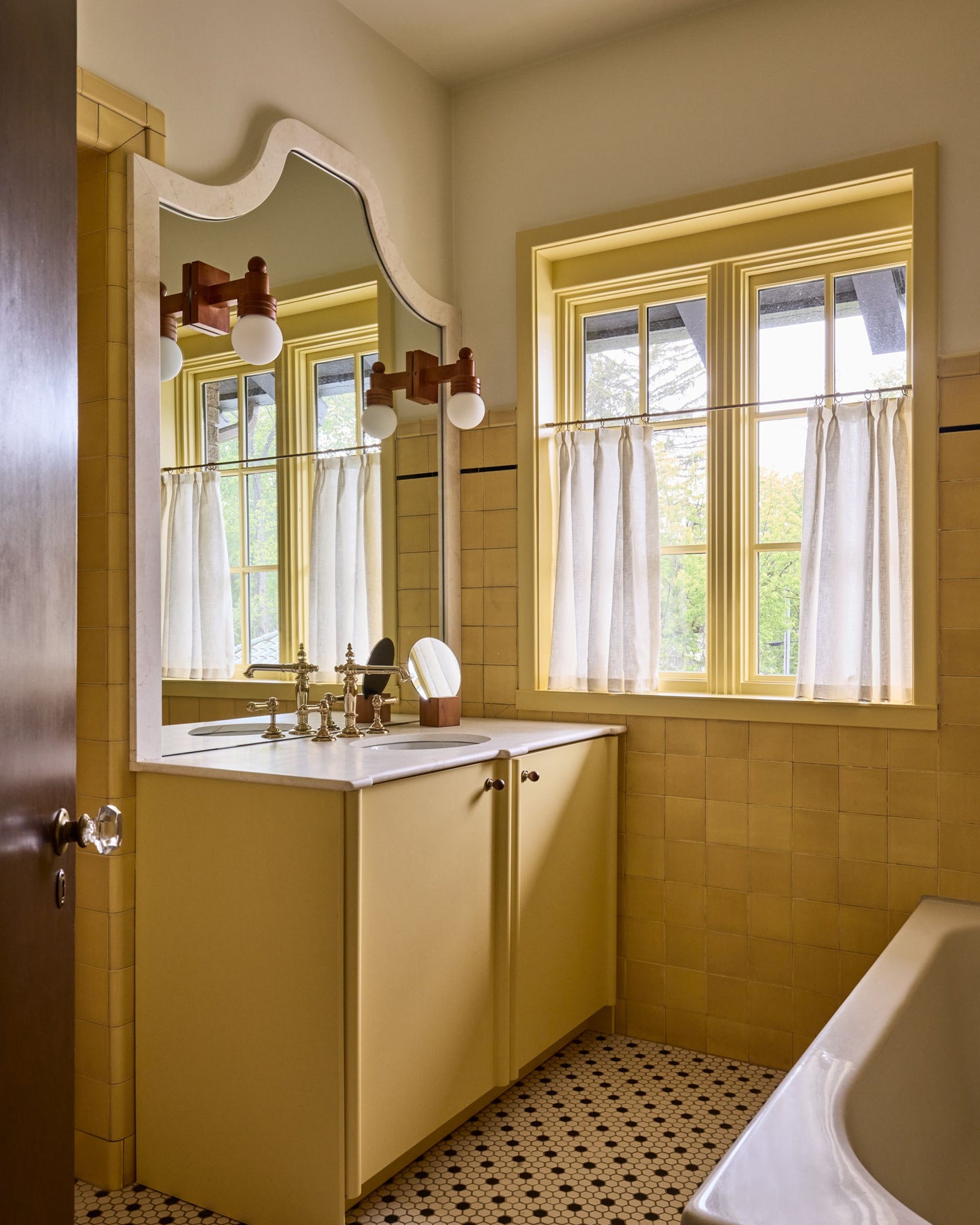 A vintage-style bathroom interior with mustard yellow tiled walls, a matching yellow vanity cabinet, a white countertop, a large arched mirror, and a window with white curtains.