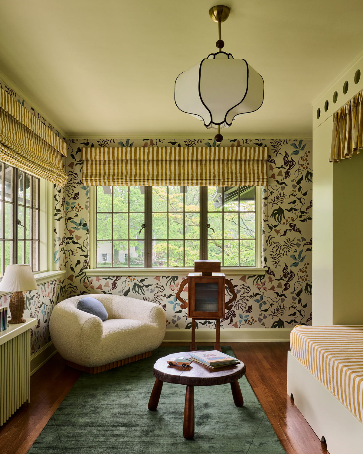 A styled interior room with floral wallpaper, striped window blinds, a cream boucle armchair, a small round wooden coffee table, a green rug, and a hanging pendant light.