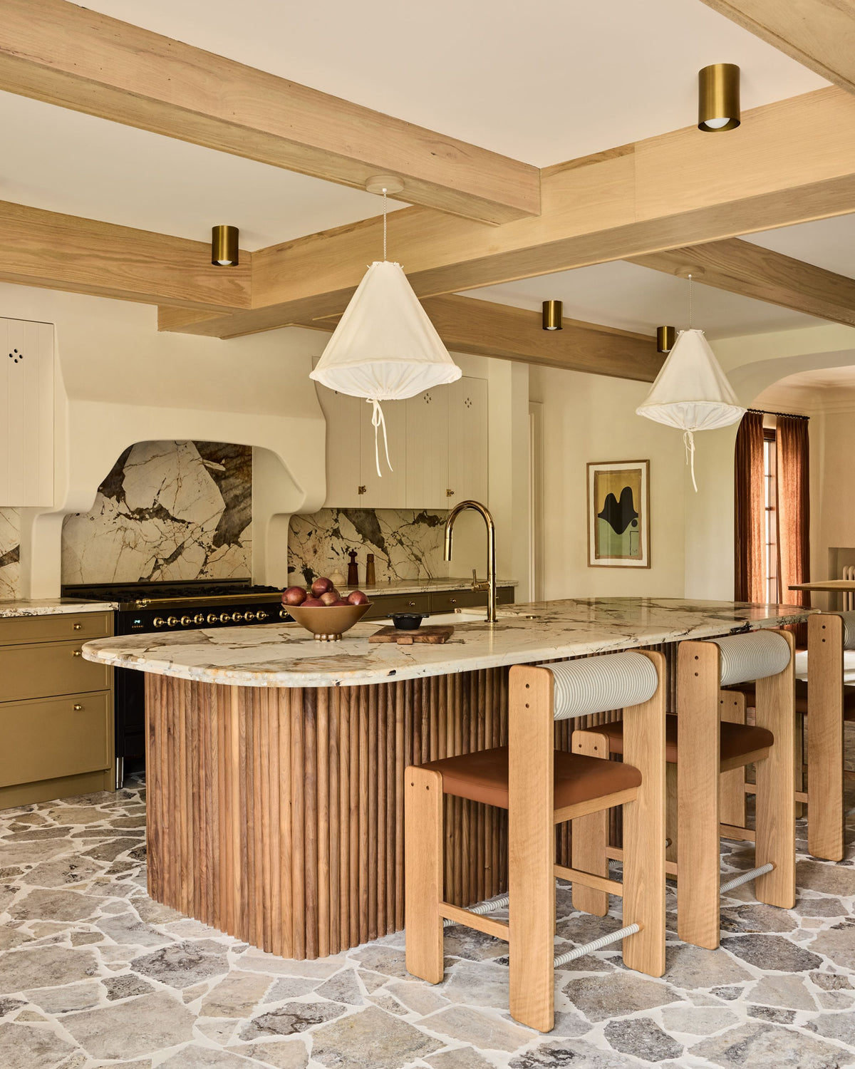 A modern kitchen island with a fluted wood base, a marble-patterned countertop, and three wooden bar stools with light cushions. The room features exposed wood ceiling beams, brass pendant lights, a stone-tiled backsplash, and a stone floor.