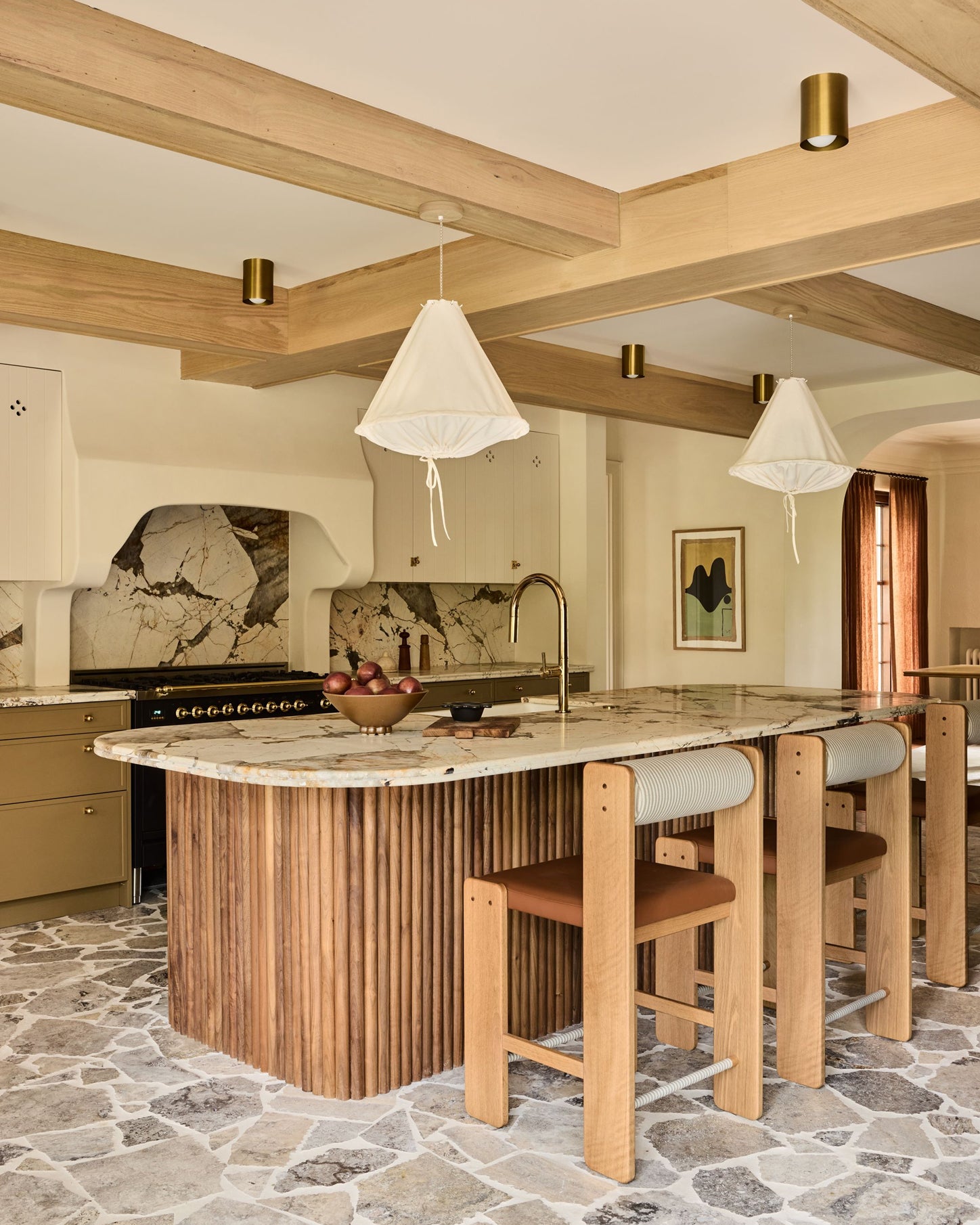 A modern kitchen island with a fluted wood base, a marble-patterned countertop, and three wooden bar stools with light cushions. The room features exposed wood ceiling beams, brass pendant lights, a stone-tiled backsplash, and a stone floor.