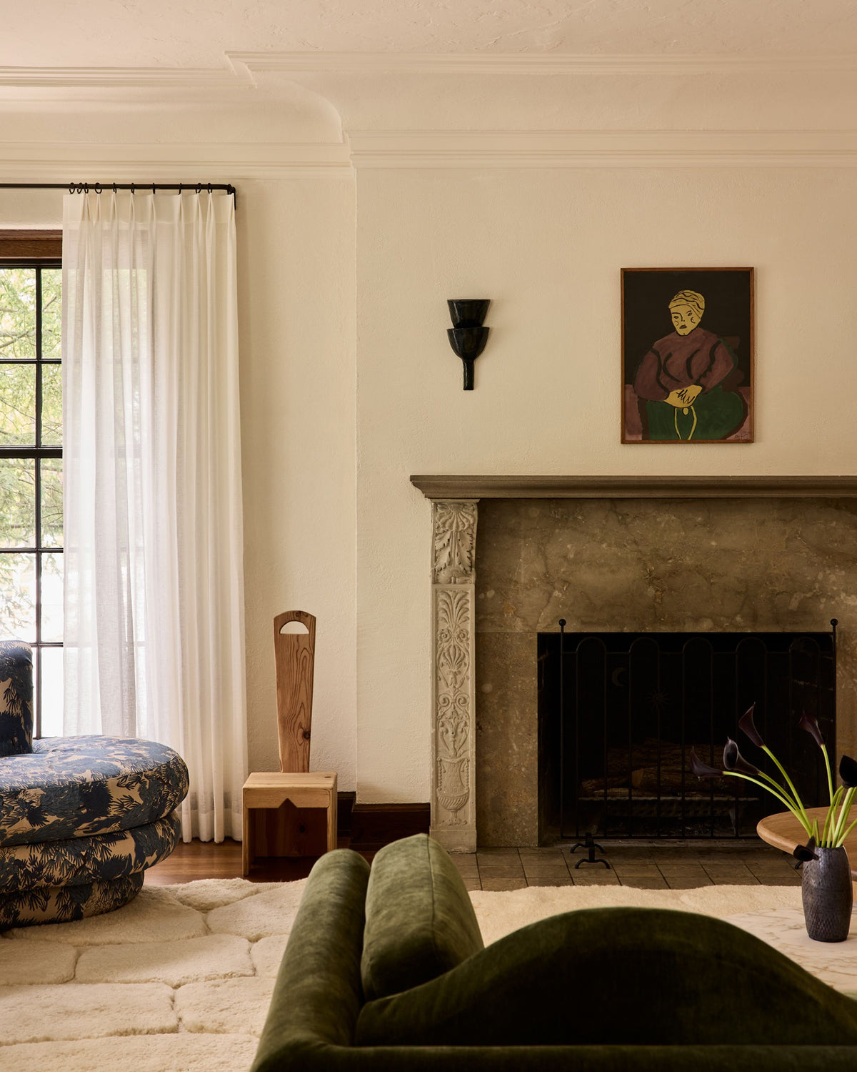 A styled interior room with a stone fireplace, cream walls, sheer white curtains, a dark green velvet armchair in the foreground, a blue floral ottoman on the left, and a framed portrait hanging above the fireplace.