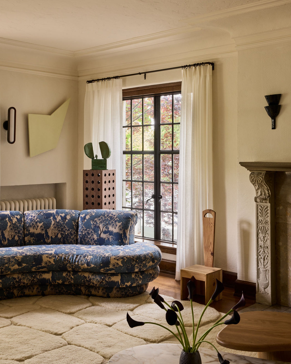 A styled interior room featuring a curved blue and beige patterned sofa, a large window with white curtains, a fireplace with a carved mantel, and a textured cream rug.