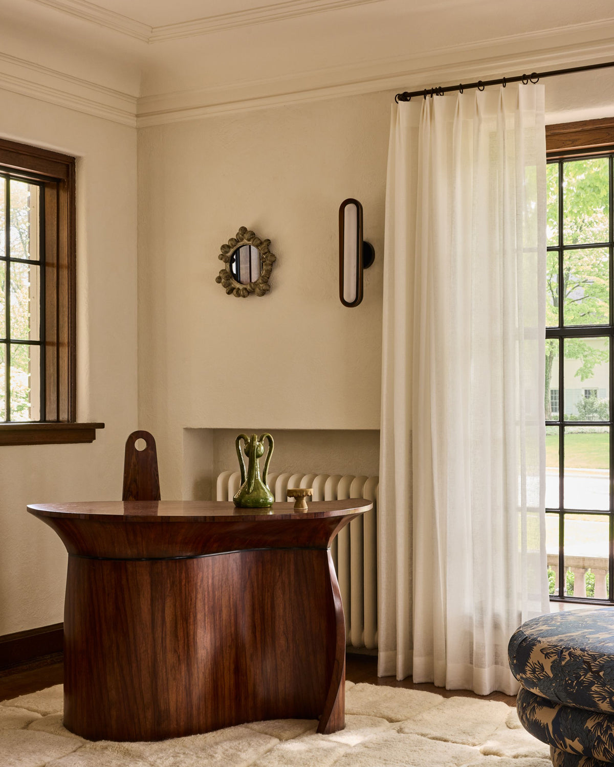A styled interior room featuring a dark wood writing desk as the primary focal point set against cream walls with a window, sheer white curtain, and a decorative green vase on top.