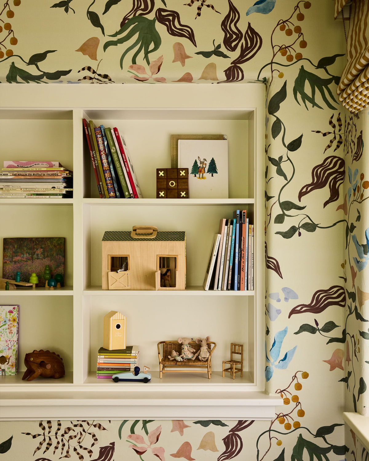 A built-in white bookshelf with cubbies styled with children's books, a wooden dollhouse, small toys, and framed art, set against a wall covered in a botanical patterned wallpaper.