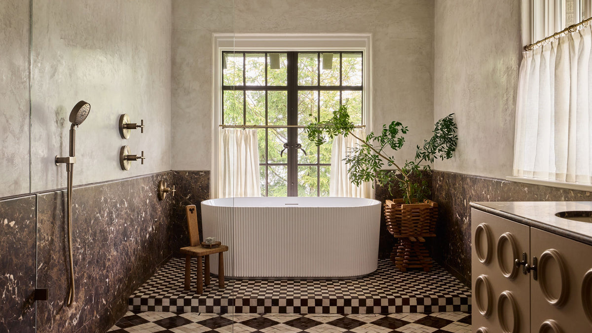 A modern freestanding oval bathtub in a styled bathroom with textured walls, checkered floor, a window with sheer curtains, and potted plants.