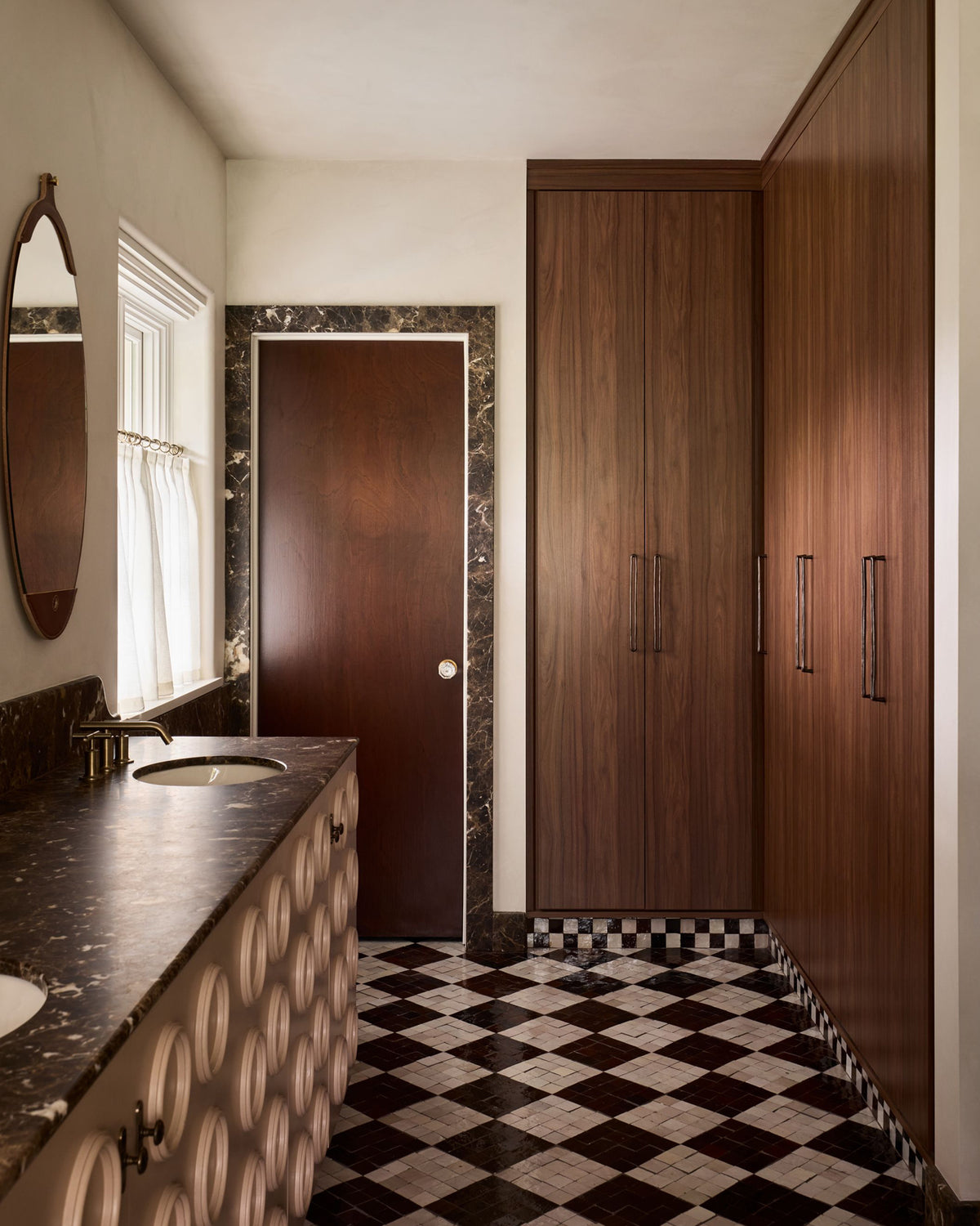 A bathroom interior with dark wood cabinetry, a checkered black-and-white tile floor, a marble countertop with a sink, and a dark wood door framed by marble trim.