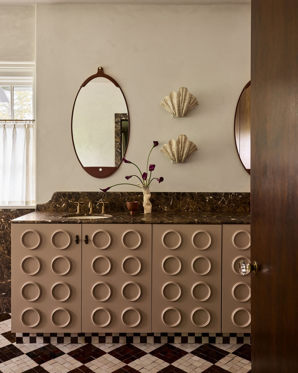 A bathroom vanity featuring beige cabinets with a raised circular pattern, a dark marble countertop, an oval mirror with a dark wood frame, and two shell-shaped wall decor pieces on a beige wall.