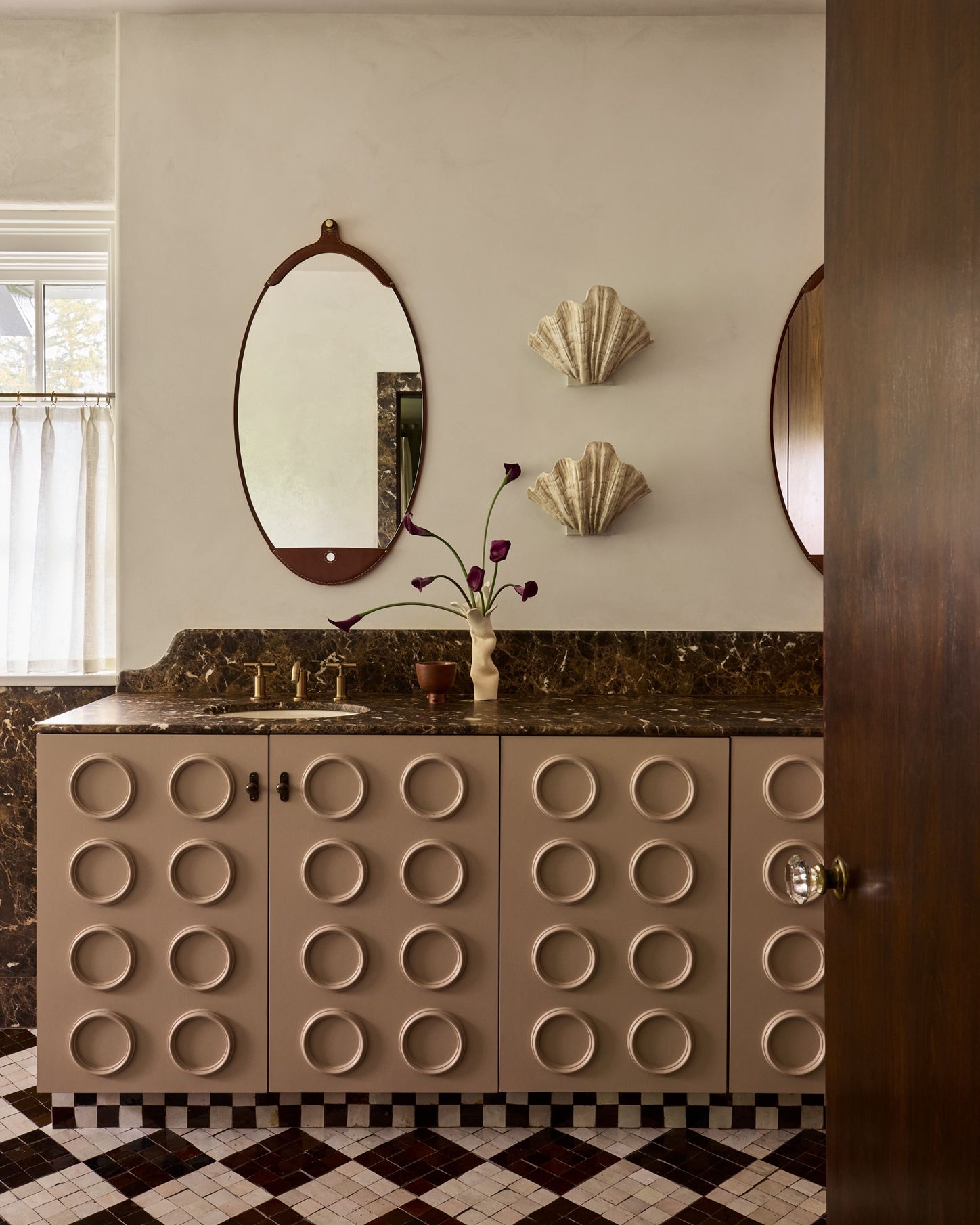 A bathroom vanity featuring beige cabinets with a raised circular pattern, a dark marble countertop, an oval mirror with a dark wood frame, and two shell-shaped wall decor pieces on a beige wall.