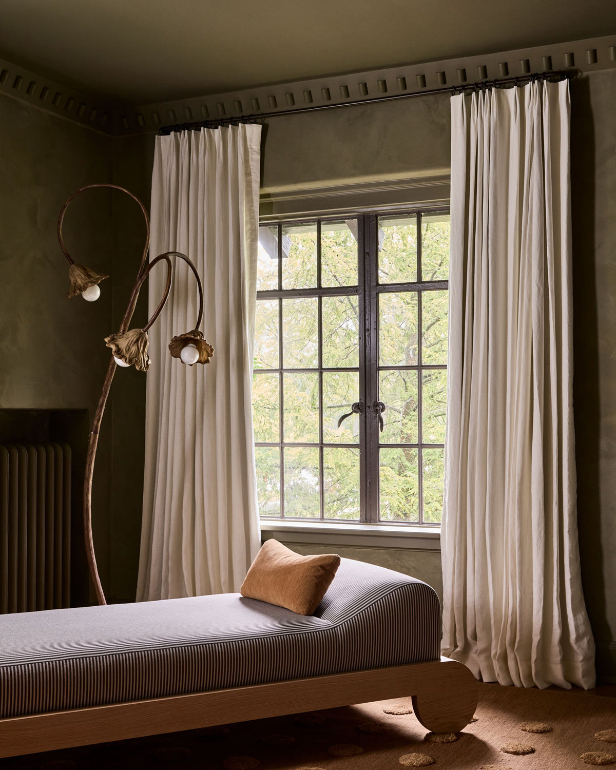 A styled interior room with a bed/chaise in the foreground, a tall sculptural floor lamp, and two long cream curtains framing a multi-pane window with visible greenery outside.