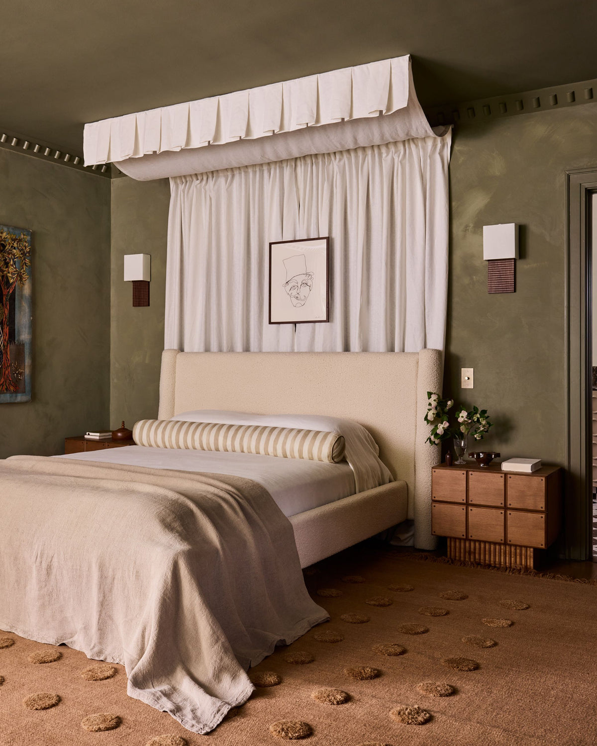 A styled bedroom featuring a bed dressed in neutral beige linens, a striped bolster pillow, a draped white canopy with a ruffled valance, olive green textured walls, and a brown patterned rug.