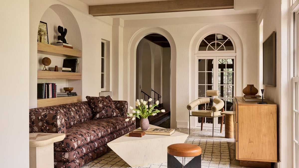 A styled interior living room with a dark floral patterned sofa, a white marble coffee table, a wooden cabinet, and arched architectural details in the background.