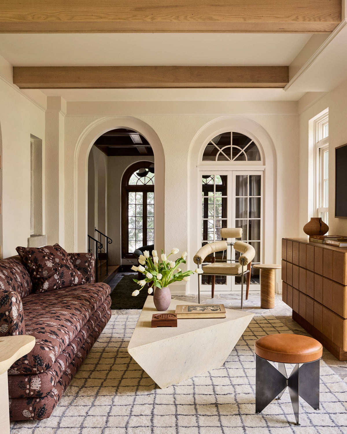 A styled living room interior featuring a brown floral patterned sofa, a white geometric coffee table with a vase of white tulips, a patterned rug, a wooden dresser, and arched windows in the background.
