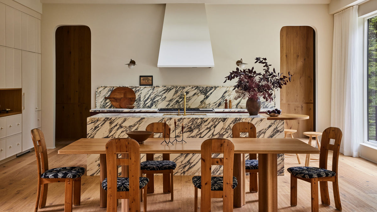 A modern kitchen and dining area featuring a light wood dining table and six matching wooden chairs with patterned seat cushions, set against a marble-tiled kitchen counter and a white range hood.