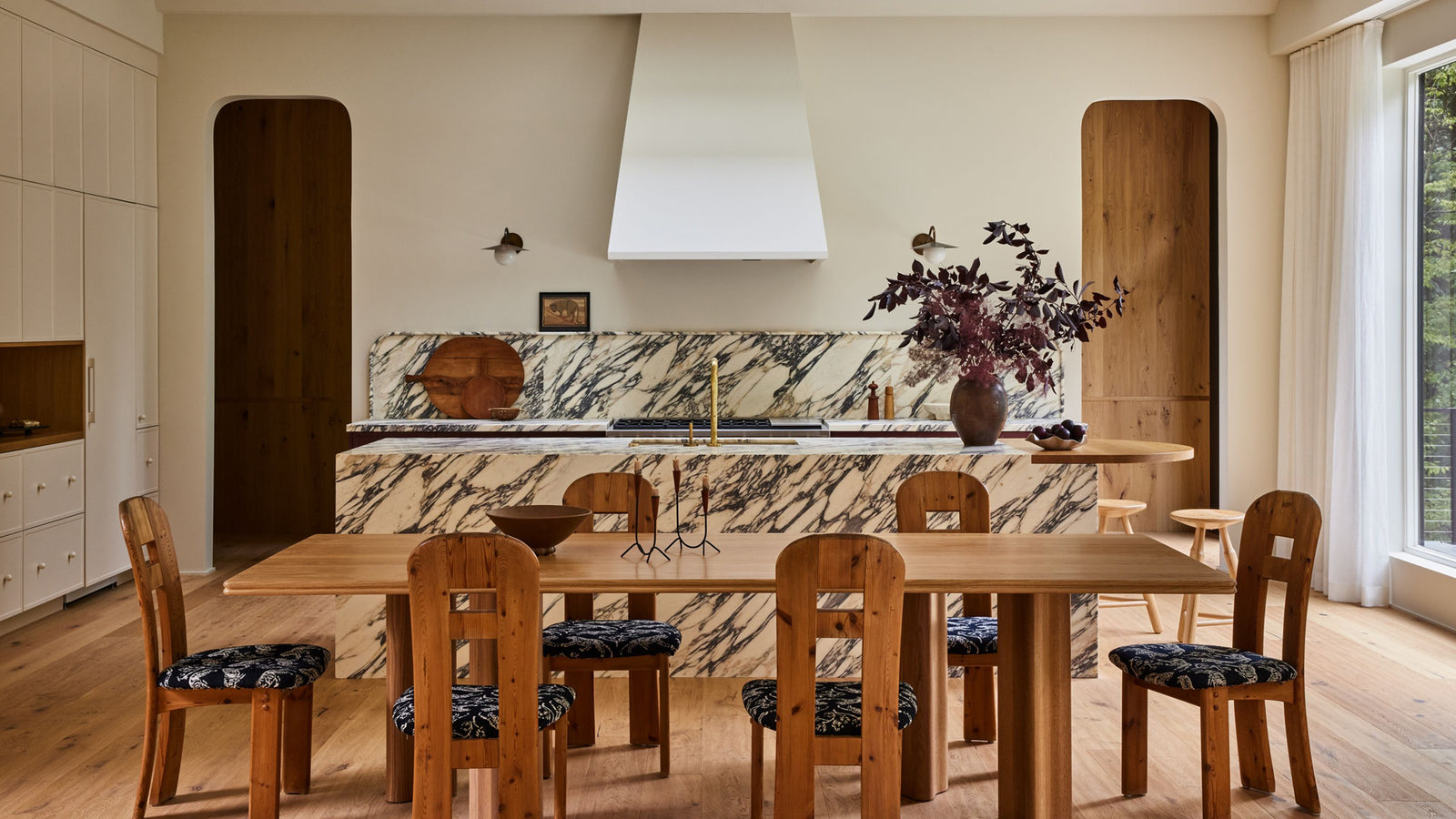 A modern kitchen and dining area featuring a light wood dining table and six matching wooden chairs with patterned seat cushions, set against a marble-tiled kitchen counter and a white range hood.