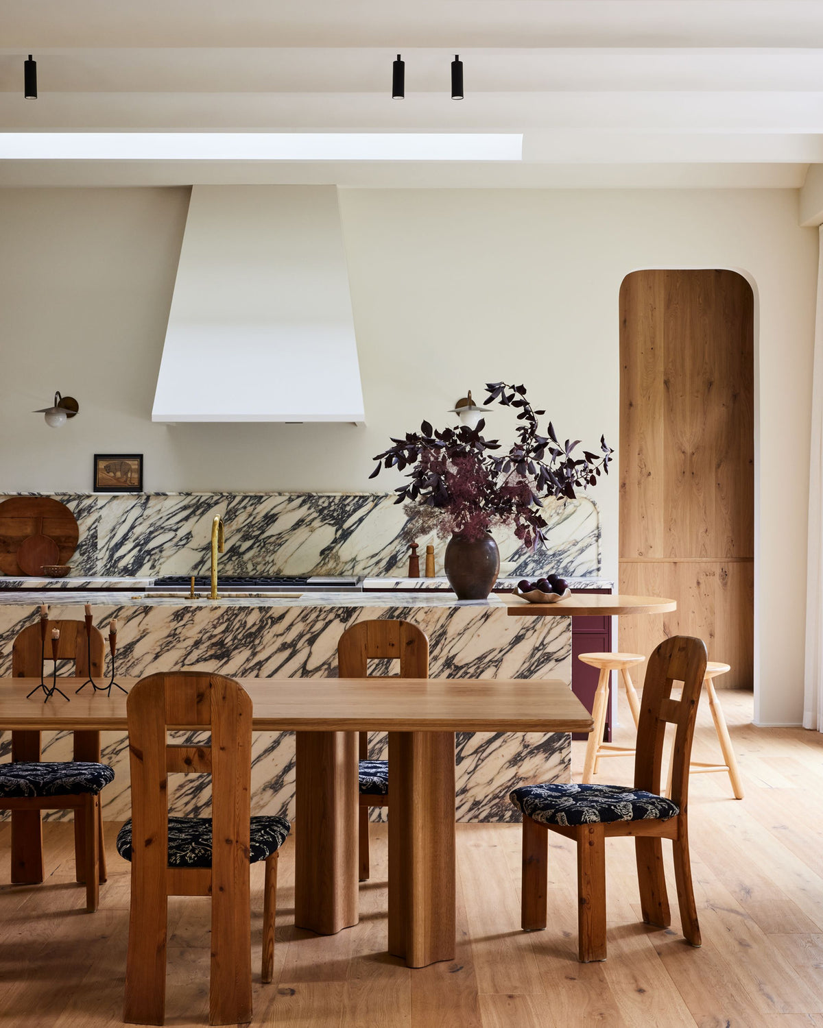 A warm, modern dining area featuring a light oak wooden dining table and matching wooden chairs with patterned seat cushions, set in a contemporary kitchen with marble backsplash and a white range hood.