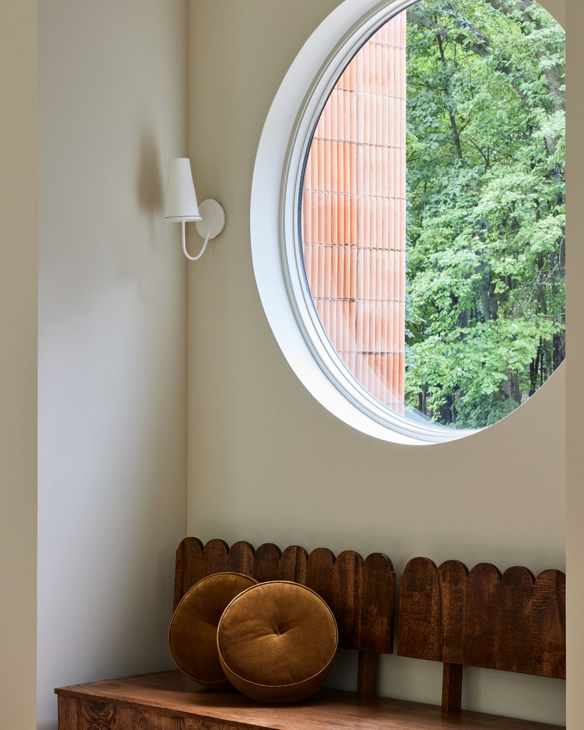 A wooden bench with a scalloped backrest and two round brown cushions, positioned beneath a large oval window showing green trees and a brick wall outside. A white wall sconce is mounted on the beige wall.