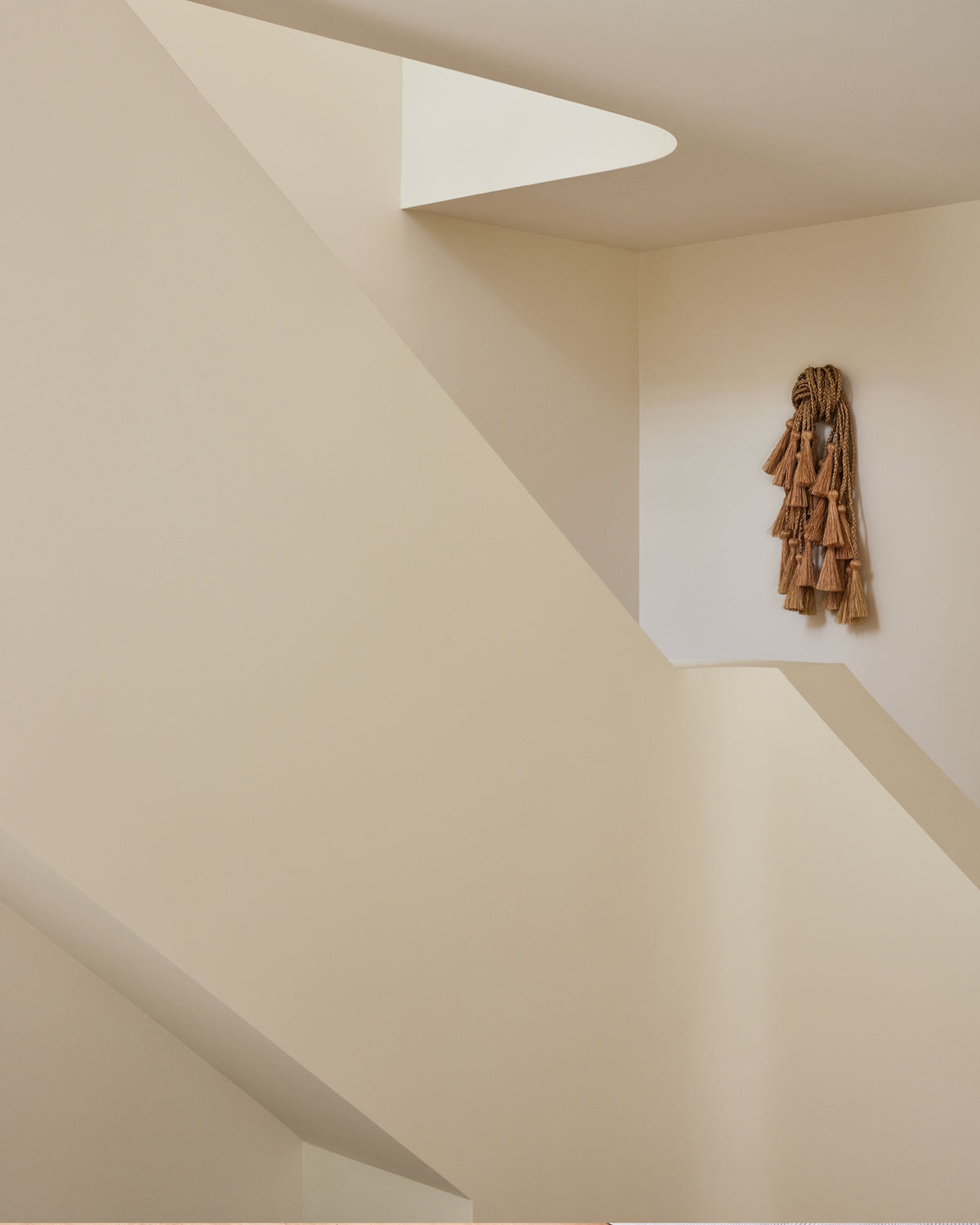 A minimalist interior scene with cream-colored walls and a staircase, featuring a small brown ruffled textile or scarf hanging on the right wall.