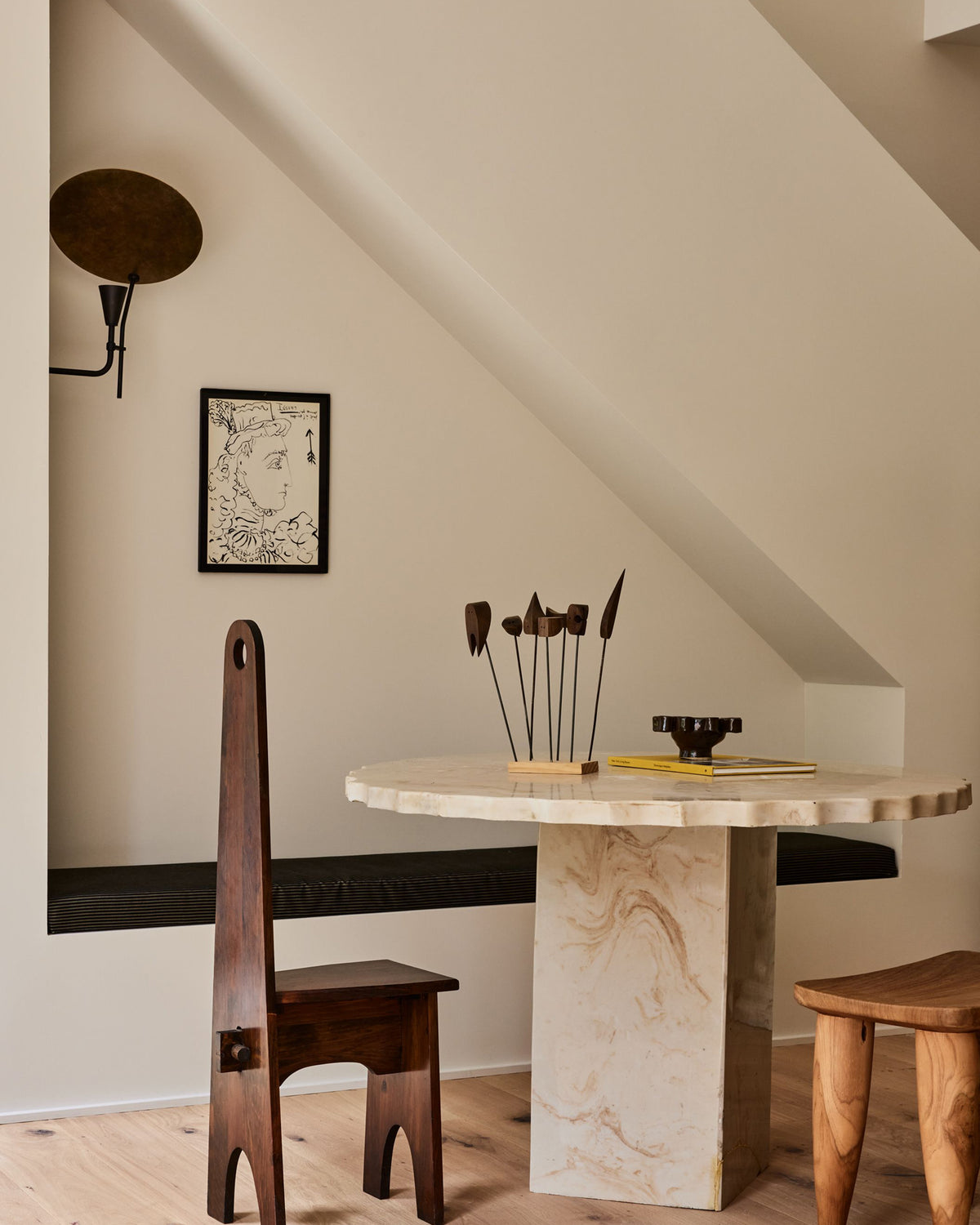 A styled interior scene with a round marble-top dining table, a tall dark wooden chair, a wooden stool, and a slanted cream wall under a ceiling.