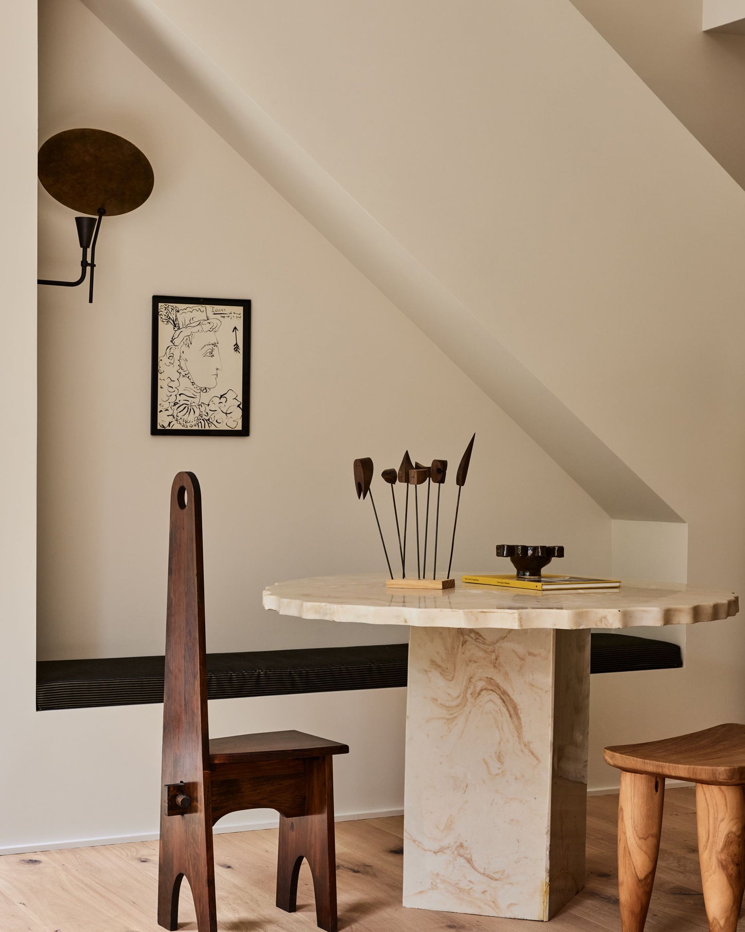 A styled interior scene with a round marble-top dining table, a tall dark wooden chair, a wooden stool, and a slanted cream wall under a ceiling.