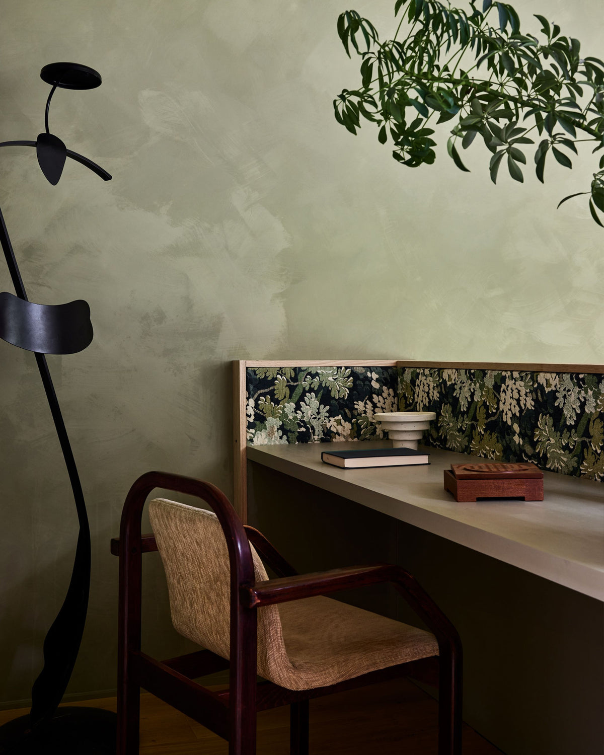 A styled interior corner featuring a wooden desk with a floral-patterned backrest, a woven seat and backrest chair, a black sculptural floor lamp, and a textured sage green wall.