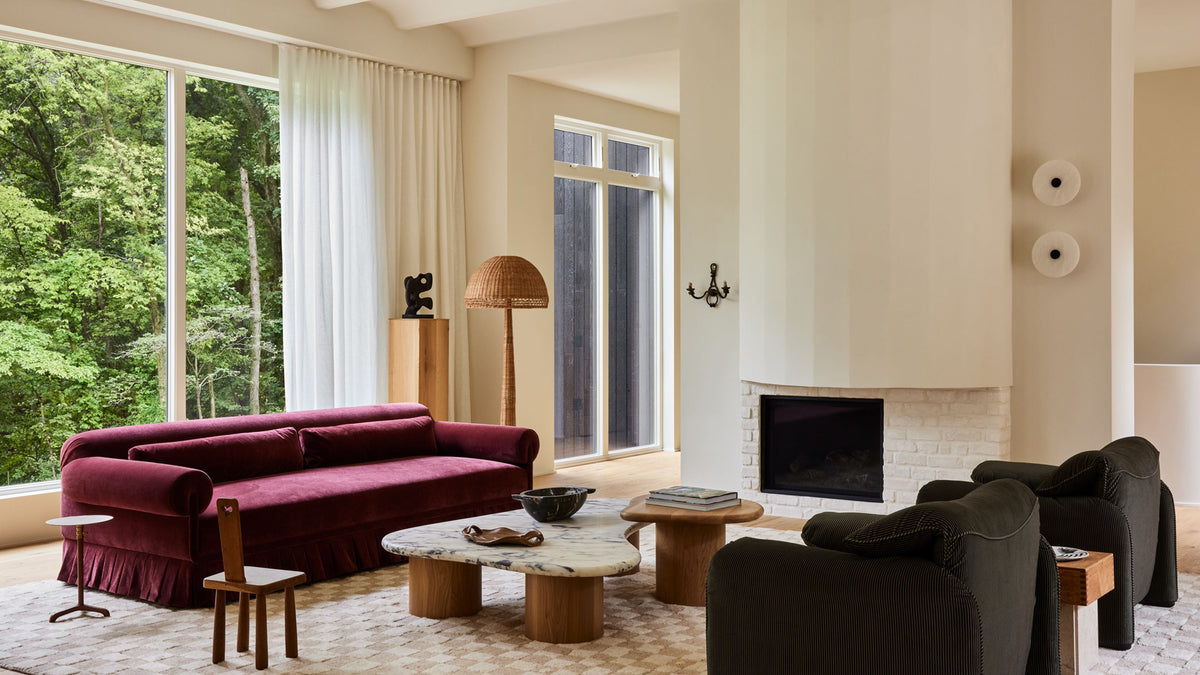 A living room featuring a prominent deep magenta velvet Alba sofa by Sarah Sherman Samuel with a skirted base, styled alongside a Skimming Stone coffee tables by Sarah Sherman Samuel, dark gray armchairs, a stone fireplace, and large windows overlooking a lush green forest. 