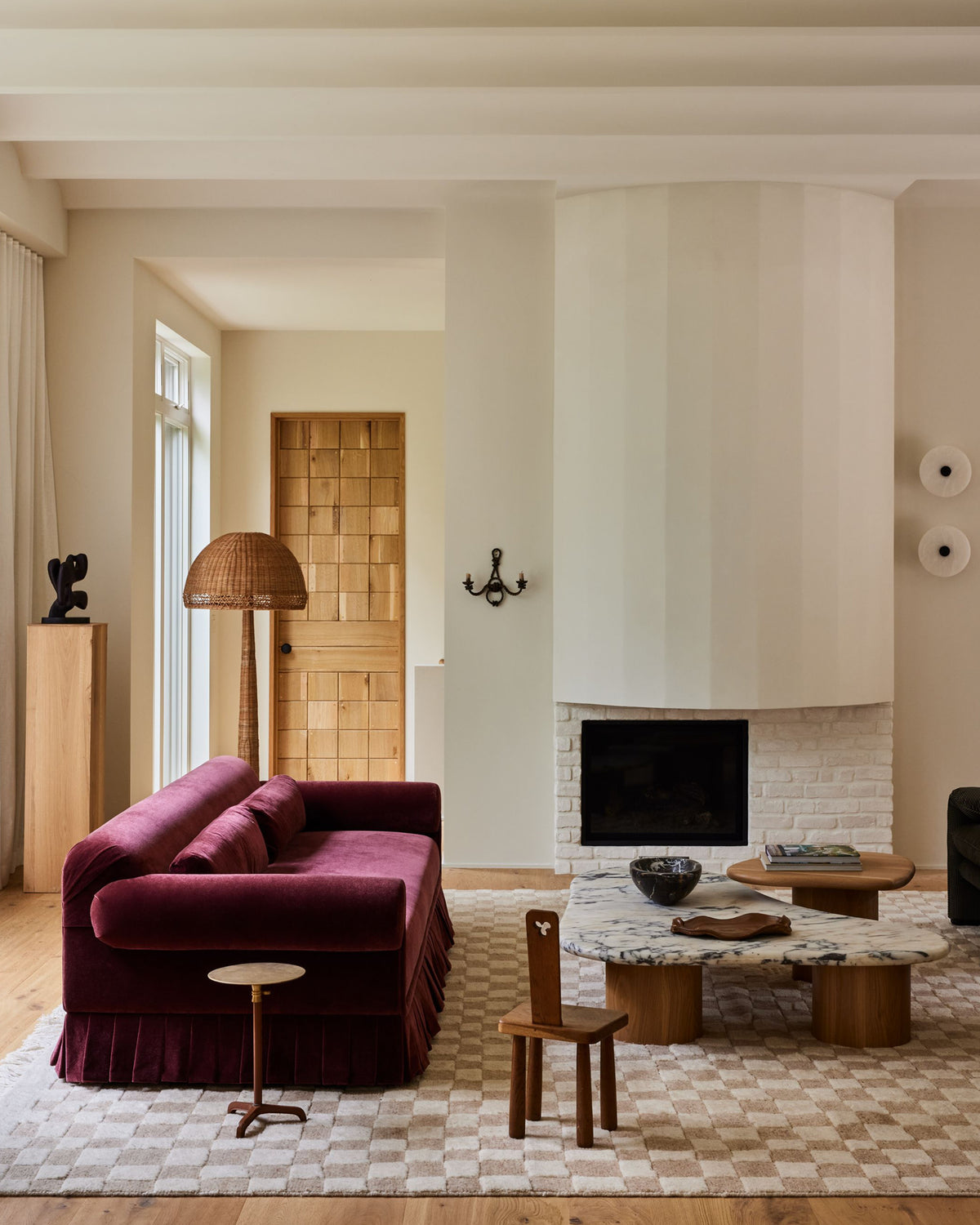 A styled living room featuring a deep burgundy velvet sofa with a pleated skirt, set on a beige and white checkered rug next to a marble-top wooden coffee table and a small wooden chair.