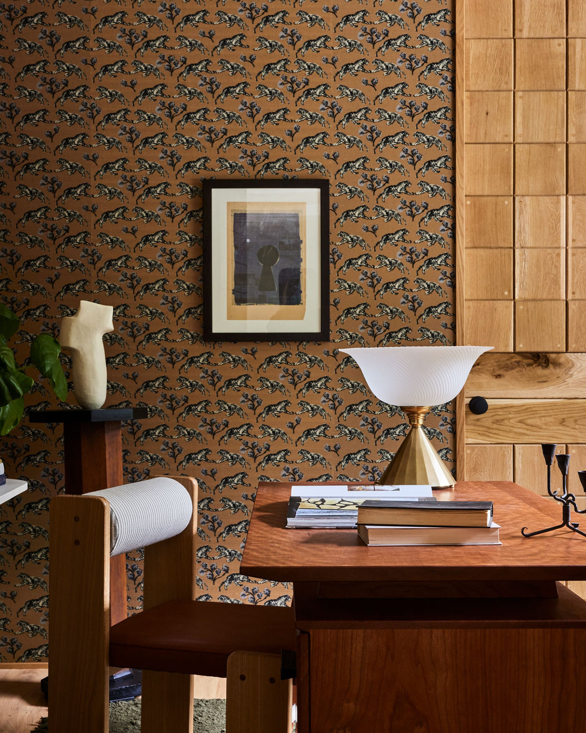 A styled interior scene featuring a brown wallpaper with a repeating fish pattern, a wooden desk with a white bowl lamp, a wooden chair, and a framed print on the wall.