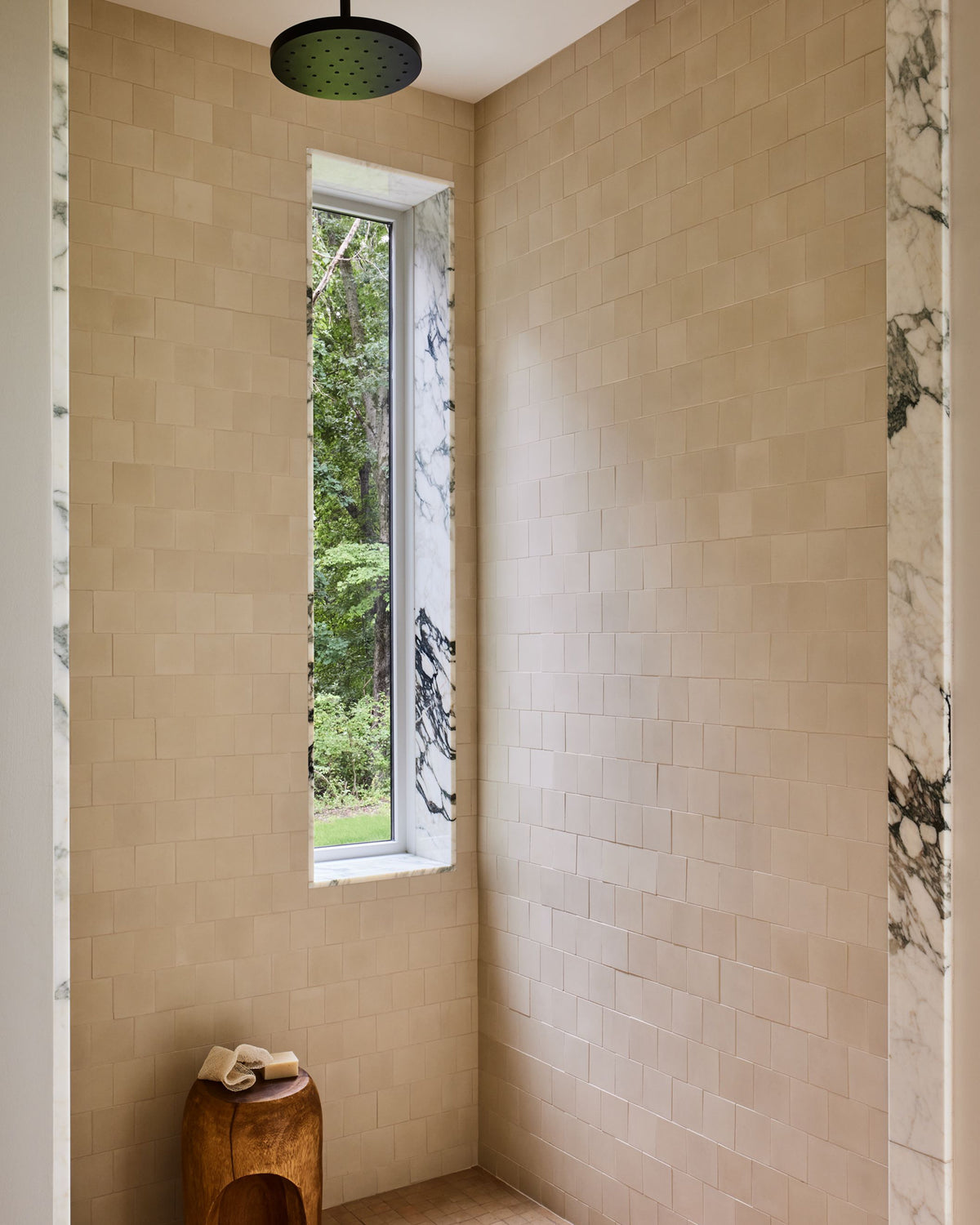A bathroom corner with beige rectangular wall tiles, a narrow vertical window showing greenery outside, a dark round ceiling shower head, and a small wooden stool with folded towels.