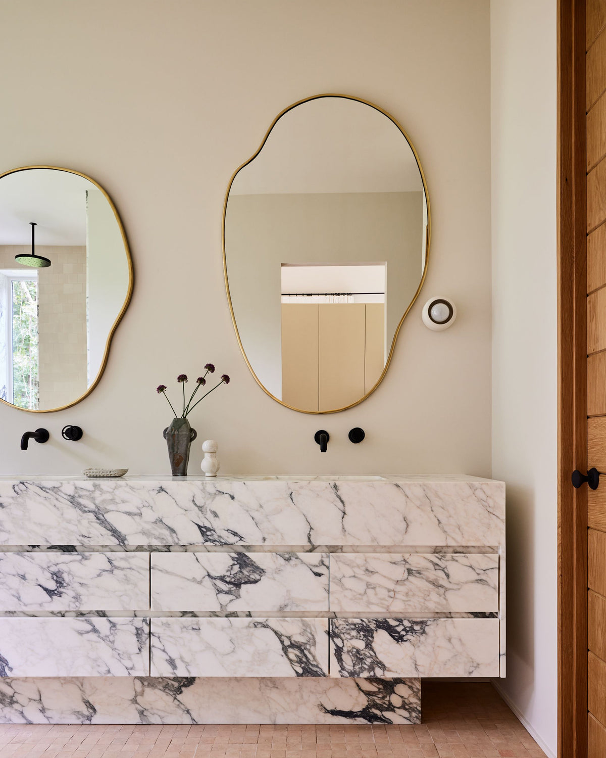 A modern bathroom vanity with a white marble front featuring dramatic black veining, paired with two large organic-shaped gold-framed mirrors on a beige wall.
