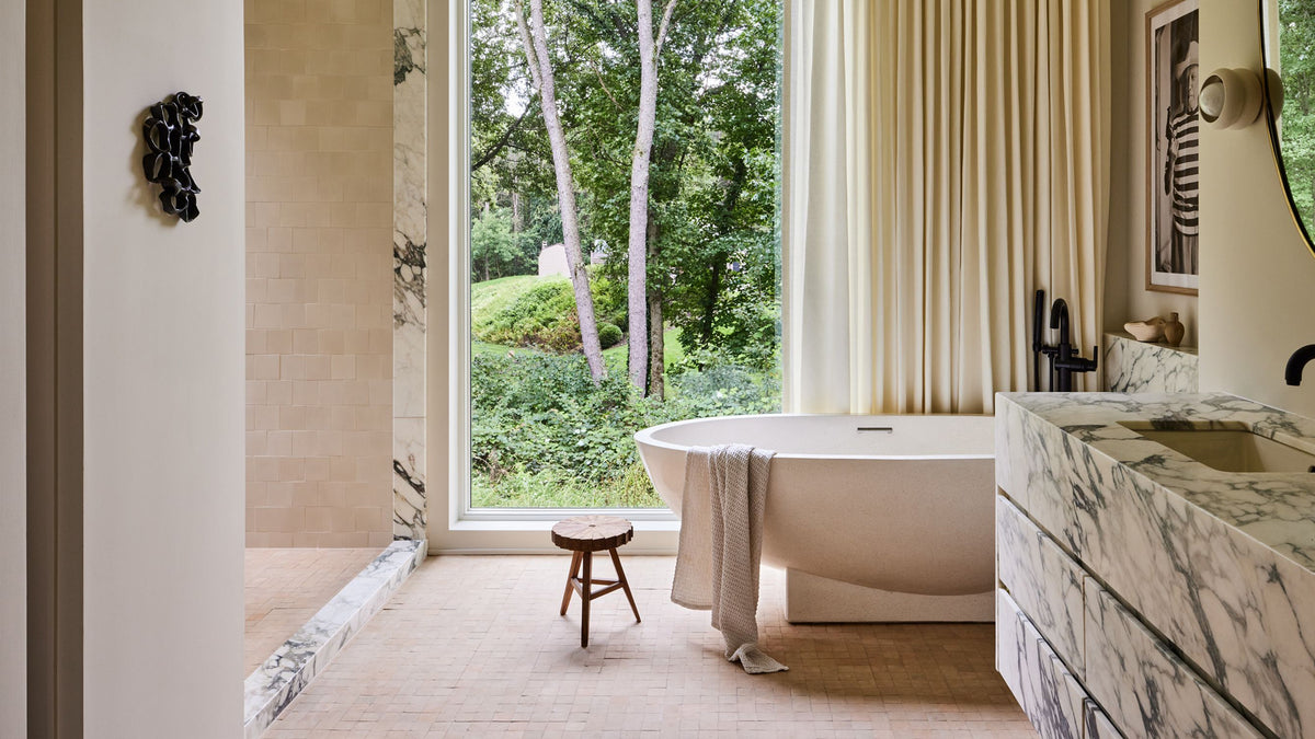 A luxury modern bathroom featuring a freestanding oval bathtub with a draped towel, a marble vanity with a sink, and a large window looking out onto a lush green garden. The room is styled with beige walls, cream curtains, and a small wooden stool.