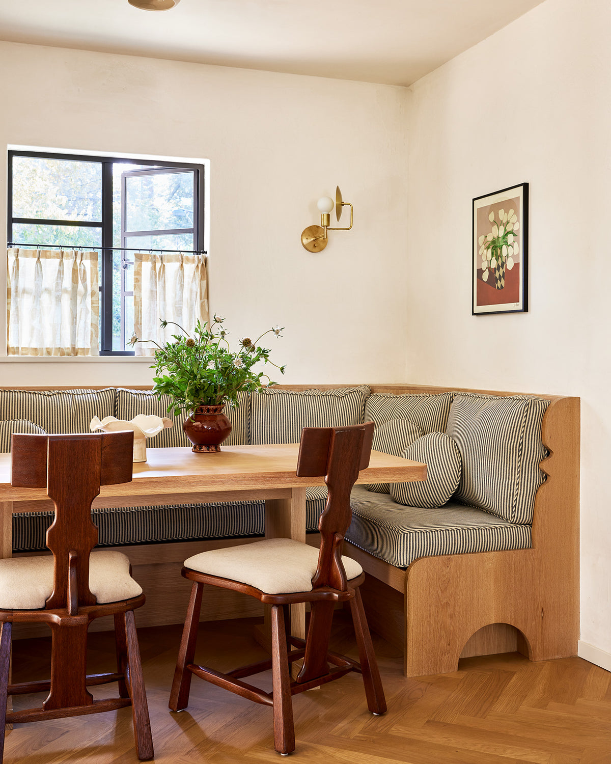 A cozy corner dining nook with a light wood table, two wooden chairs, and a built-in bench with striped cushions, set in a warm beige room with a window and wall sconce.