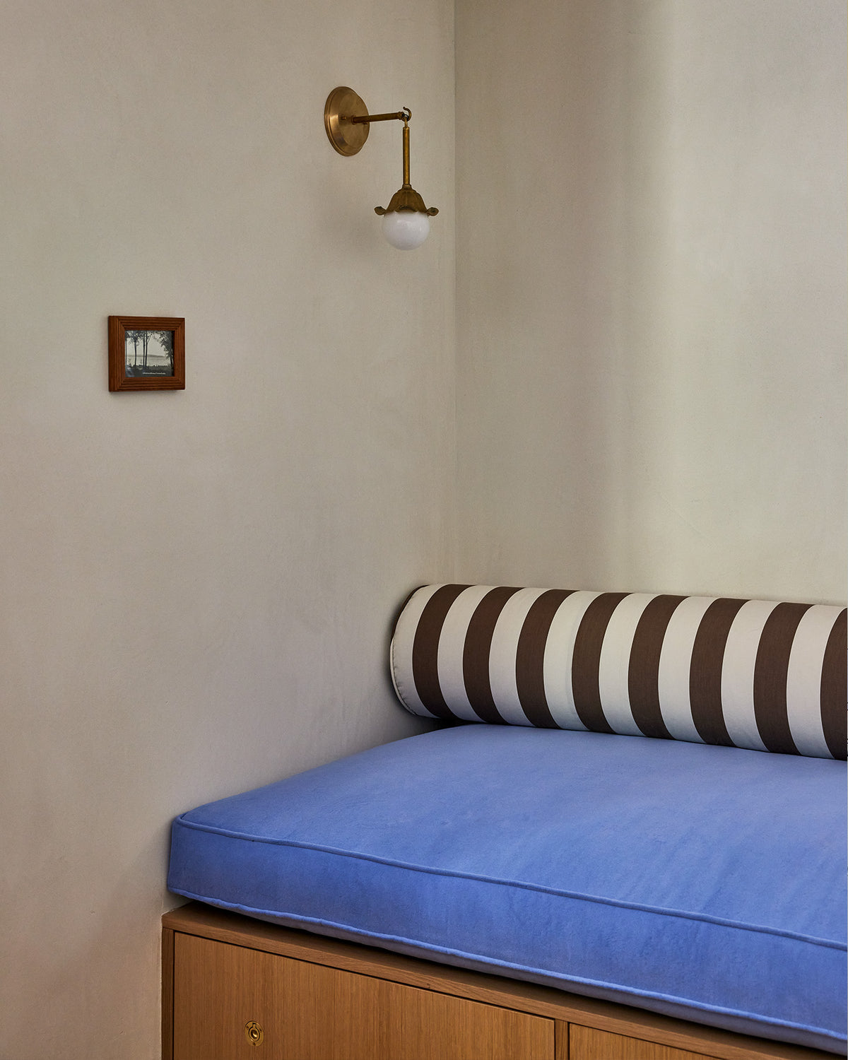 A cozy corner seating nook with a blue cushioned bench on a wooden base, a brown and white striped cylindrical pillow, and a brass wall sconce on the beige plaster wall.