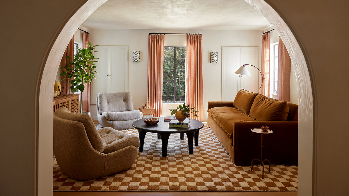 A warm, elegant living room viewed through a large arched doorway. The room features a brown leather sofa, a beige armchair, a light gray armchair, a round black coffee table, peach curtains, and a checkered tile floor.