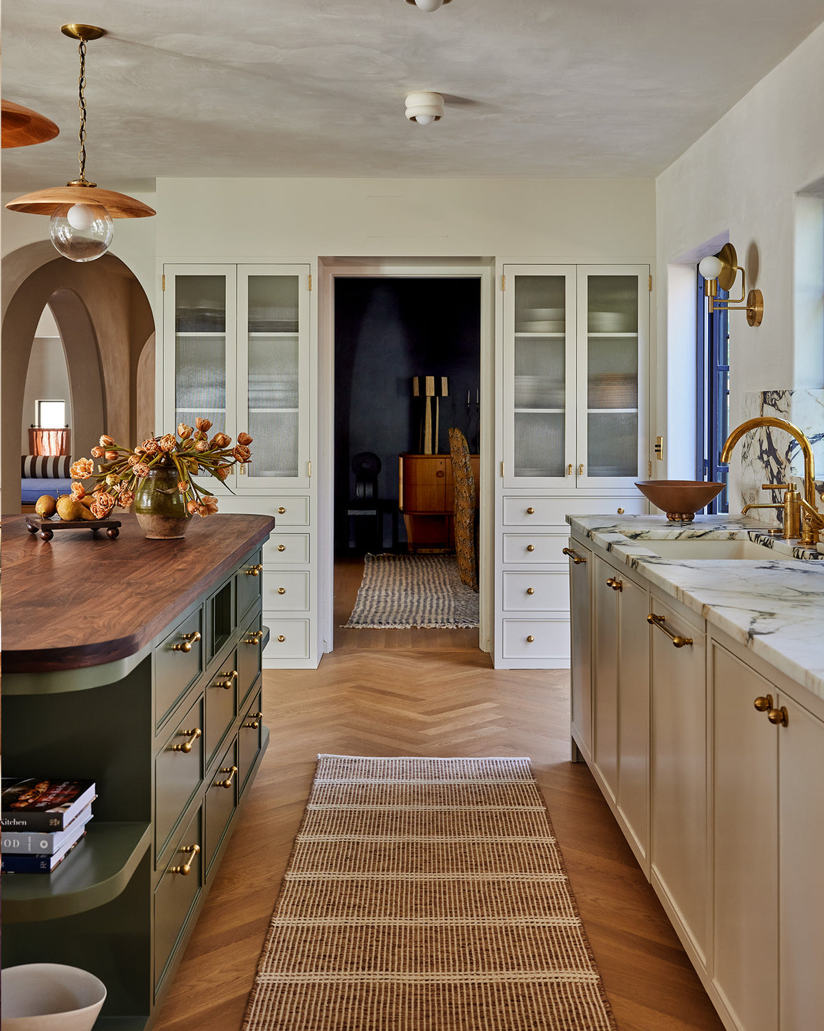 A wide interior kitchen scene with cream cabinetry, a dark green island, wood countertops, a woven rug, and brass fixtures. No person or animal is present, but this is still not a clean isolated product shot; it shows a real environment, so subject_type is Lifestyle.
