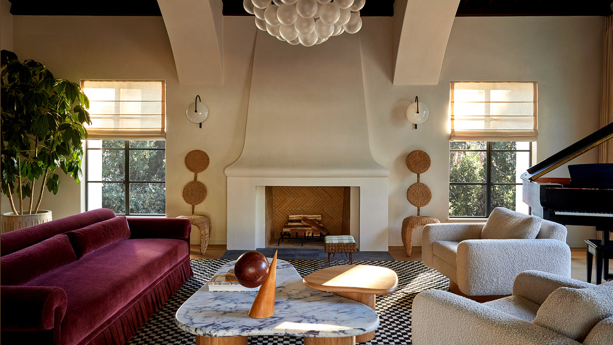 A living room interior featuring a deep burgundy velvet sofa on the left, cream armchairs on the right, a marble-top coffee table in the foreground, a white fireplace with a decorative mantel, and a black grand piano near a window on the right.
