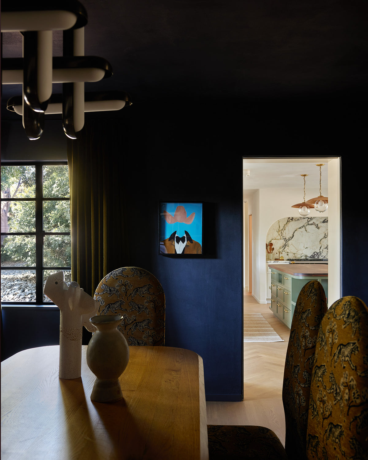 A moody dining room interior with a wooden table, patterned dining chairs, a modern multi-arm chandelier, and a small colorful wall art piece mounted on a dark blue wall. An open doorway reveals a brightly lit adjacent kitchen area.