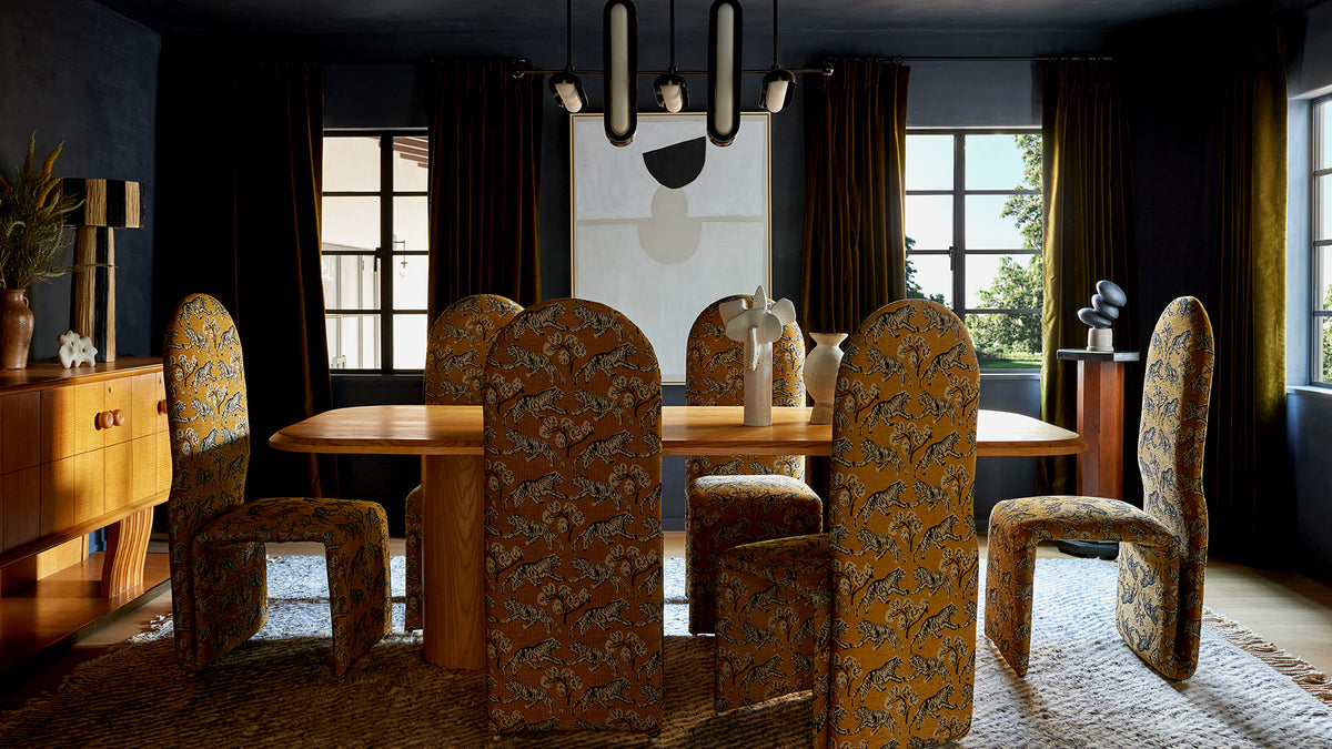 A dining room set featuring a light wood dining table and six upholstered chairs with a beige and brown floral pattern, set in a dark, elegant room with dark curtains and a modern chandelier.