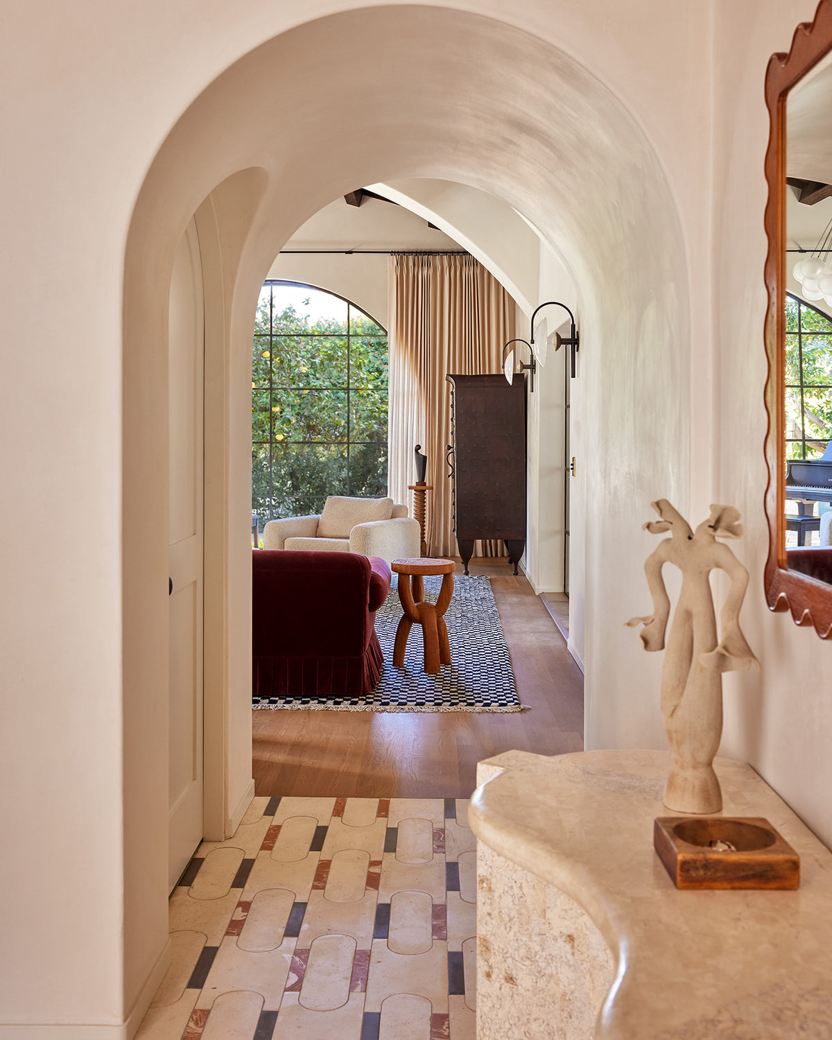 An interior architectural scene viewed through a large arched doorway, showing a furnished living room with a red sofa, wooden stool, patterned rug, tall curtains, and a marble console table in the foreground.
