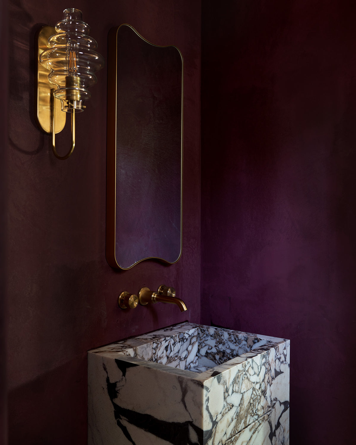 A bathroom vanity featuring a square white marble pedestal sink with black veining, a curved brass faucet, a matching wavy-edged mirror, and a gold wall sconce against a deep plum purple wall.