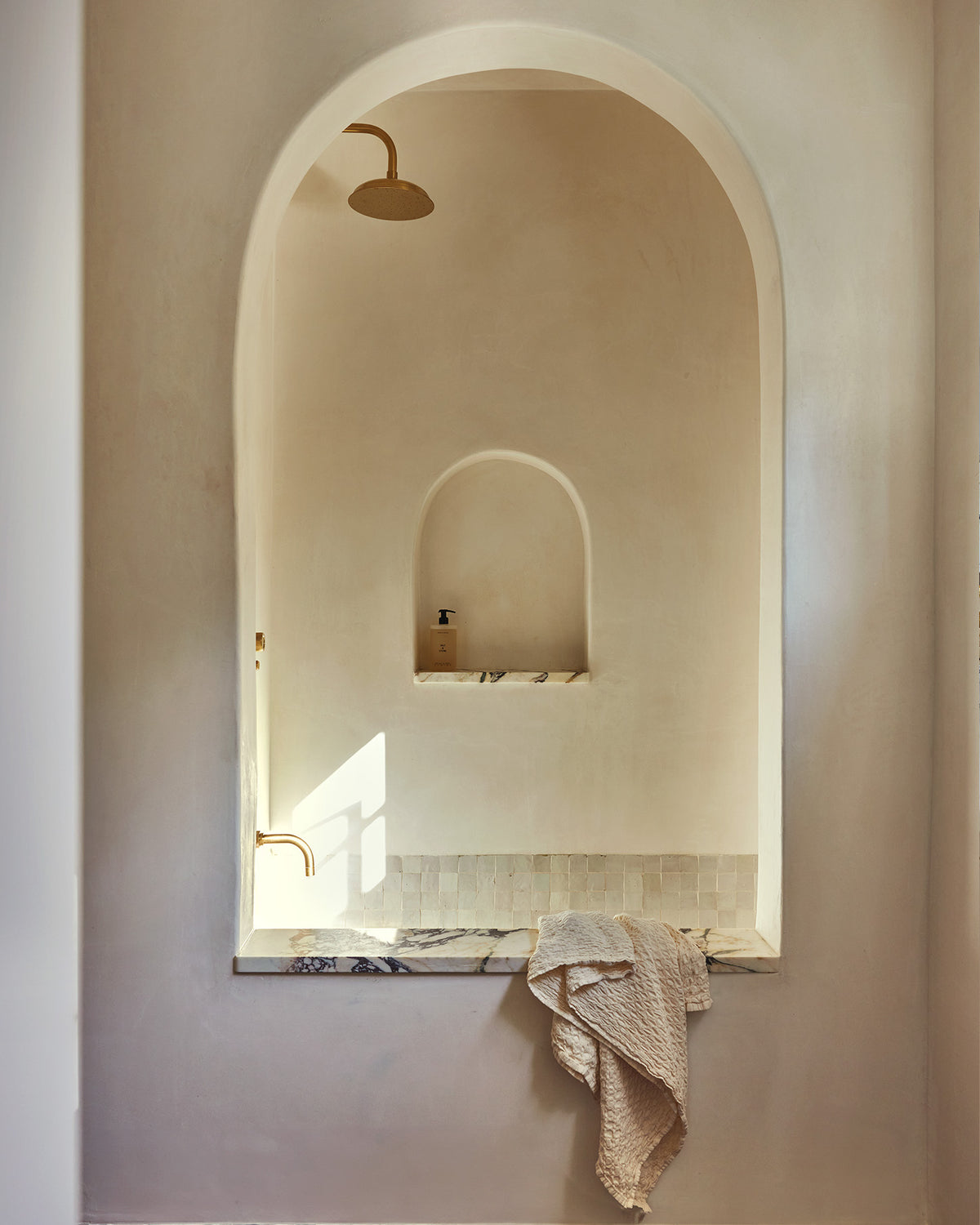 Minimalist bathroom interior featuring a large arched niche with a built-in shelf, marble ledge, draped towel, and warm brass fixtures against a soft beige plaster wall.