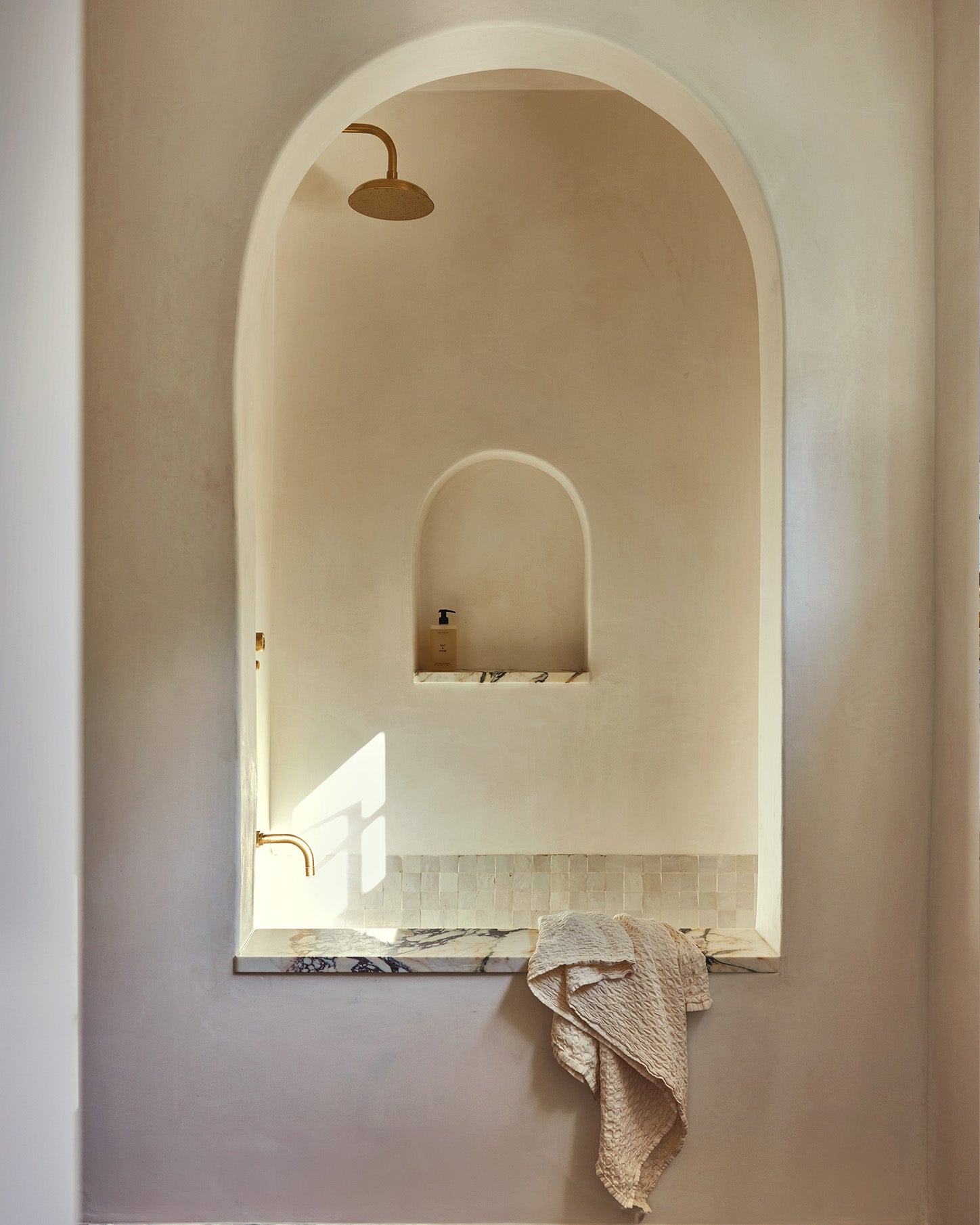 Minimalist bathroom interior featuring a large arched niche with a built-in shelf, marble ledge, draped towel, and warm brass fixtures against a soft beige plaster wall.