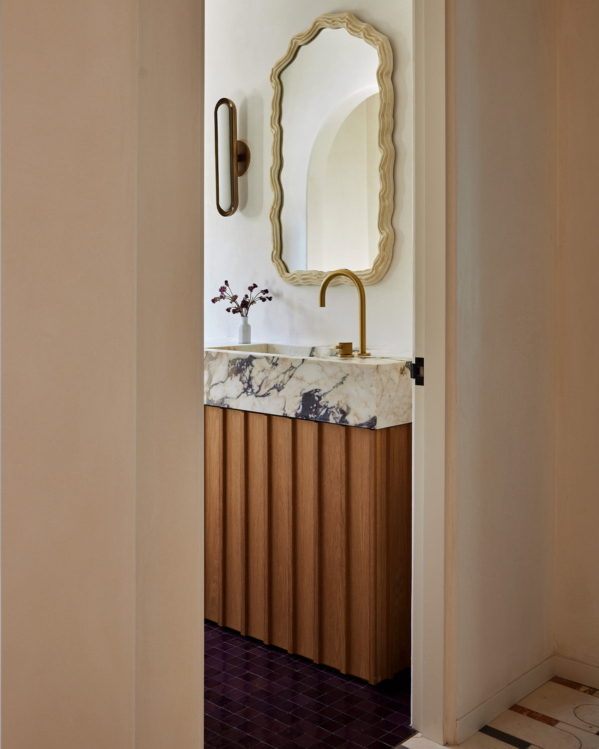 A modern bathroom vanity featuring a slatted wood cabinet, a white marble countertop with dark veining, a curved brass faucet, and a matching scalloped gold-framed mirror above.
