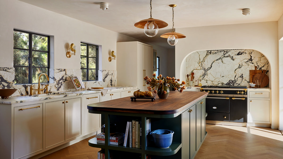 A modern kitchen interior featuring a dark green island with a rich wood countertop, white cabinets, gold hardware, and a black gas range with a white marble backsplash.