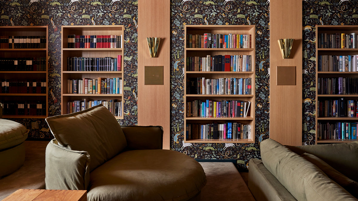 A movie room scene with tall wooden bookshelves filled with books, patterned wallpaper, wall sconces, and two upholstered lounge chairs in the foreground.