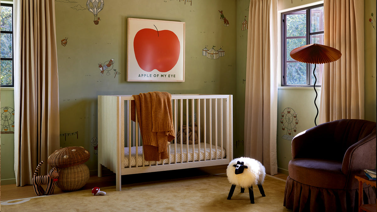 A styled nursery interior featuring a white wooden crib with a rust-colored blanket, a framed red apple artwork on the wall, beige curtains, a brown armchair, and a small sheep-shaped stool on a beige carpet.