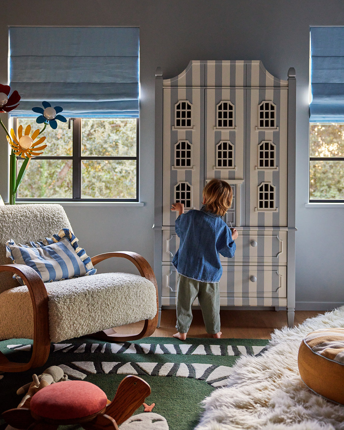 A child stands in front of a tall, narrow cabinet with vertical blue and white stripes and decorative window details, in a styled room with windows, a chair, and a rug.