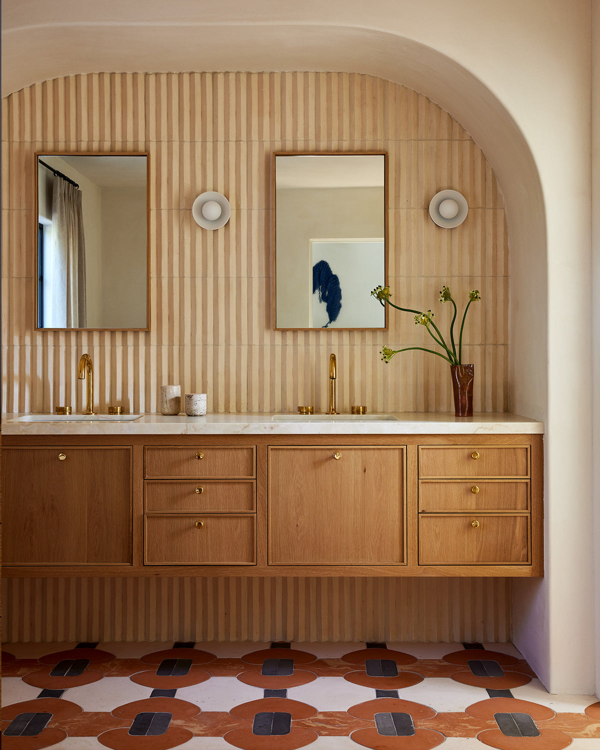 A bathroom vanity with light wood cabinets, a white countertop, two mirrors, and a ribbed beige tiled wall. A patterned terracotta and cream tile floor is visible below, and a vase with green stems sits on the counter.