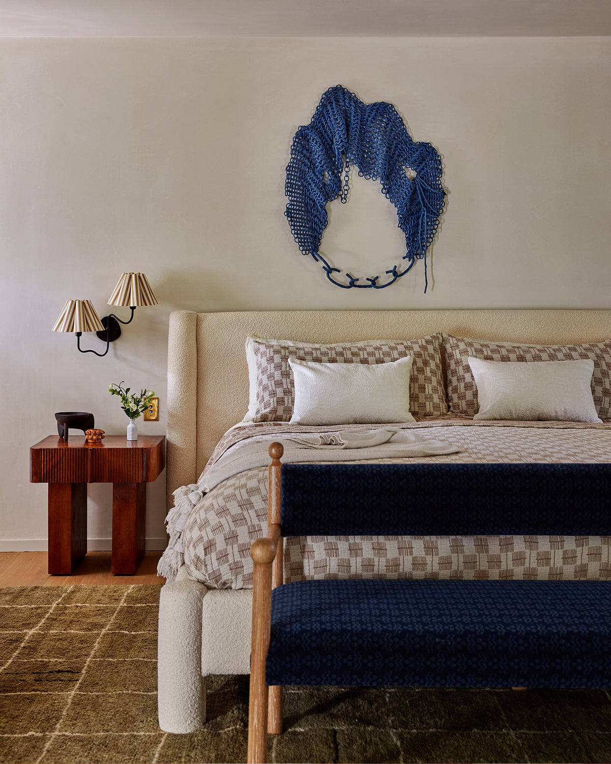 A styled bedroom scene featuring a bed with cream patterned bedding, a matching navy bench at the foot, a dark wood bedside table with two wall sconces, and a textured blue wall hanging by Taylor Kibby above the bed.