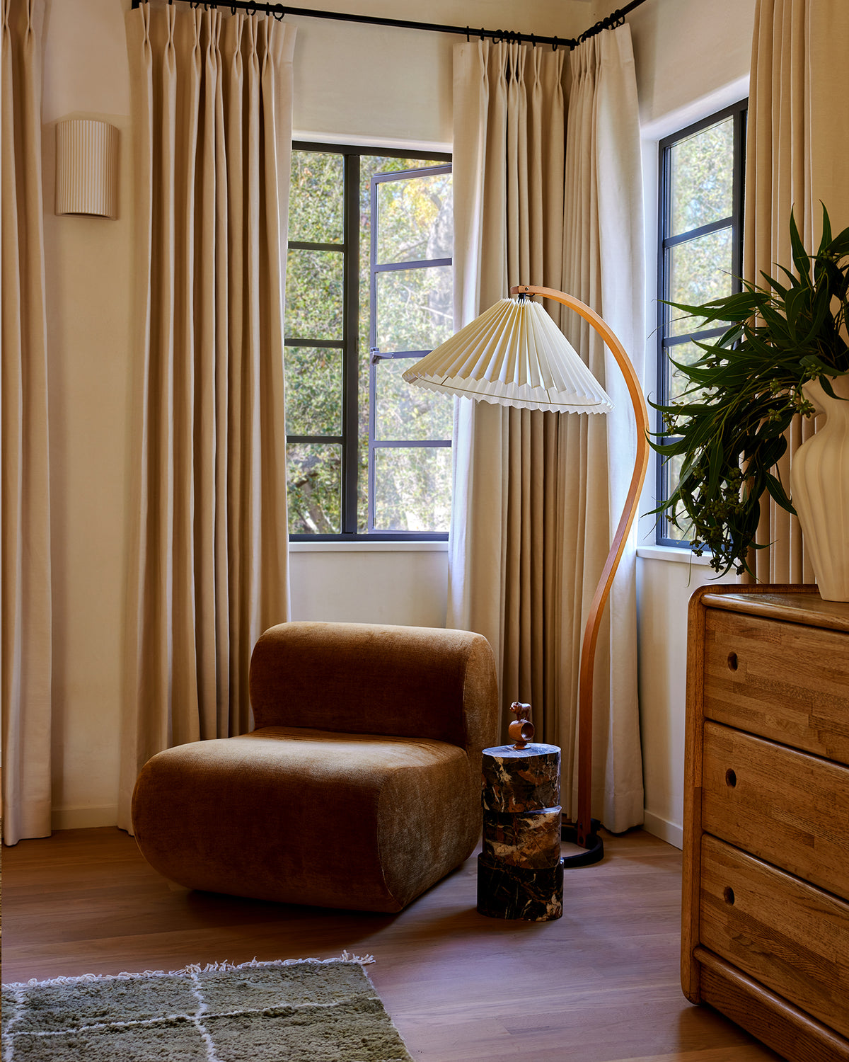 A styled corner with floor-length beige curtains, a brown upholstered lounge chair, a curved floor lamp, a wooden dresser, and a potted plant near tall windows.